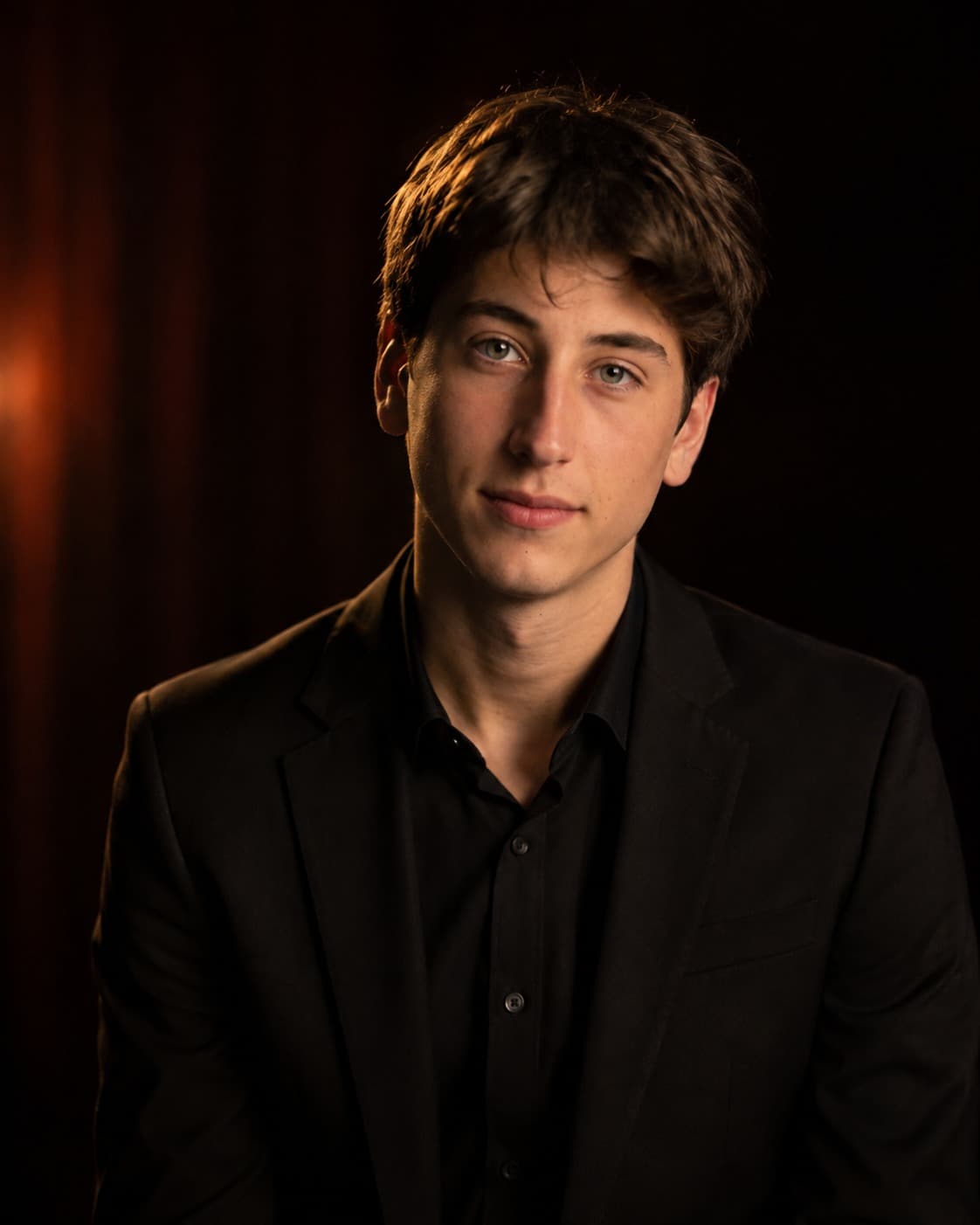 Jonah Shapiro in a black suit and black shirt, lit from one side against a dark red stage curtain